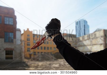 crime scene, murder and killing concept - close up of criminal or murderer hand in leather glove with blood on knife over city ruins background (staged photo)