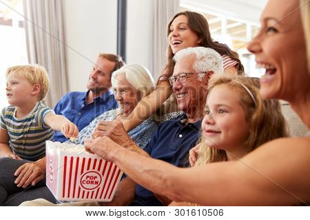 Multi-Generation Family Sitting On Sofa At Home Eating Popcorn And Watching Movie Together
