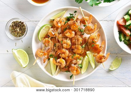 Prawns Skewers With Greens, Spices, Lime And Sauce On White Plate Over Wooden Table. Tasty Seafood. 