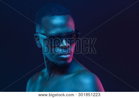 Close-up Portrait Of African Man Standing In Sunglasses And With Naked Shoulders In Neon Light