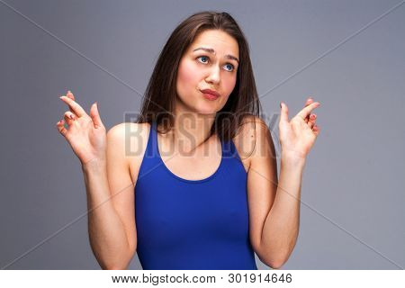 Body language and emotions concept. Glad dark haired beautiful satisfied girl beggs God to help her, keeps fingers crossed, wishes good luck, prays indoor isolated over gray studio background.