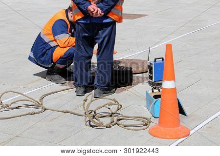 Workers In Uniform Stand Over The Open Sewer Hatch On A Street. Concept Of Repair Of Sewage, Undergr