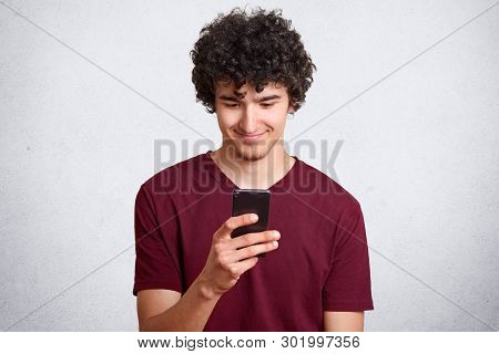 Portrait Of Pleased Attractive Man With Curly Hair And Little Beard Holding Smartphone And Staring A