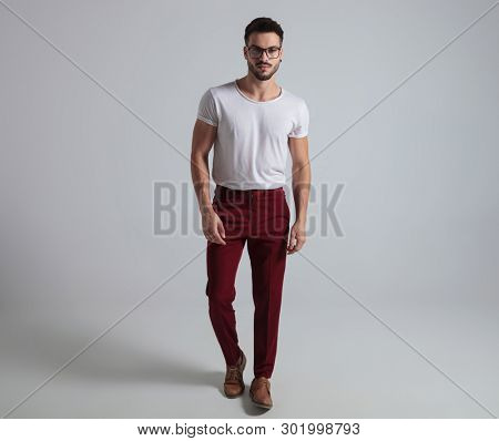 Confident guy stepping forward while wearing glasses, a white t-shirt and red pants on gray studio background