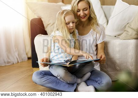 Happy Family. Blonde Young Mother Reading A Book To Her Cute Daughter While Sitting At Wooden Floor 