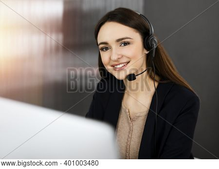 Young Friendly Girl In Headsets Is Talking To A Firms Client, While Sitting At The Desk In Sunny Off