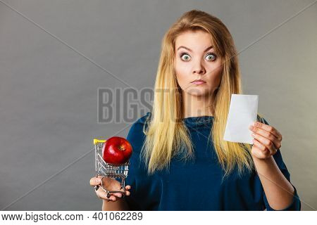 Worried Woman Holding Shopping Basket With Fruits Looking At Bill Receipt Being Scared Of Huge Price