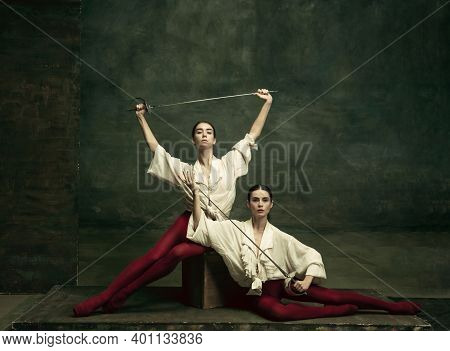 Lovely. Two Young Female Ballet Dancers Like Duelists With Swords On Dark Green Background. Caucasia