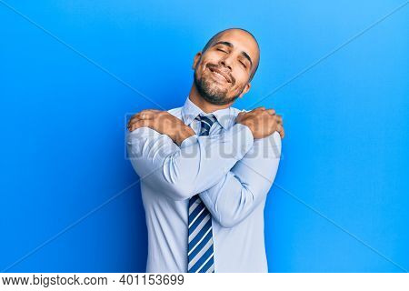 Hispanic adult man wearing business shirt and tie hugging oneself happy and positive, smiling confident. self love and self care 