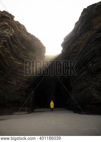 Person In Yellow Jacket Standing Infront Of Massive Rock Wall Cove Formation At Praia As Catedrais C