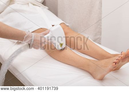 Process Of Laser Hair Removal Of Leg Hair. Young Woman In Cosmetic Robe In Salon Lies On Couch. Beau