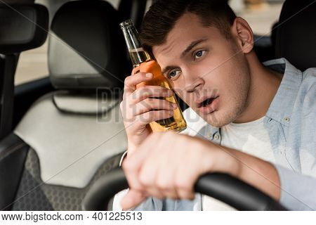 Drunk Man Holding Bottle Of Alcohol Near Head While Driving Car On Blurred Foreground.