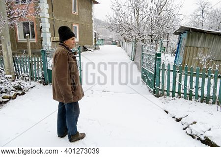 Elderly Man Standing On The Rural Street Near The Iron Gates And House During A Snowfall. Concept Of