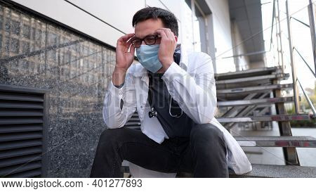 Worried And Tired Doctor Sitting On Stairs Outside Hospital. Exhausted Physician In White Lab Coat A