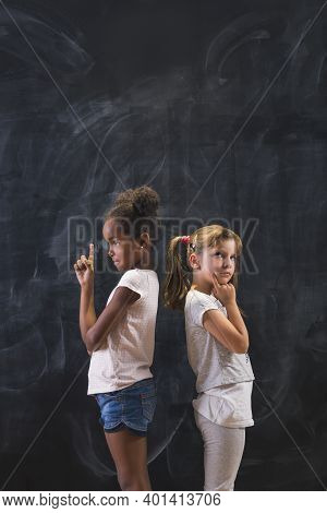 Two Beautiful Schoolgirls Standing In Front Of A Chalkboard Back To Back, Pensive And Serious, Think