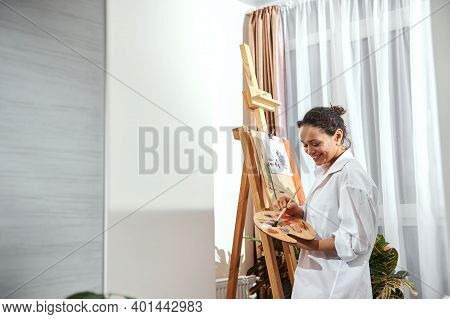 View Of A Beautiful Female Painter With Gathered Hair In A Bun And Paint Brushes In Her Hair Standin