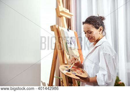 Side View Of A Smiling Happy Female Painter Mixing Oil Paint On A Palette And Standing In Front Of A