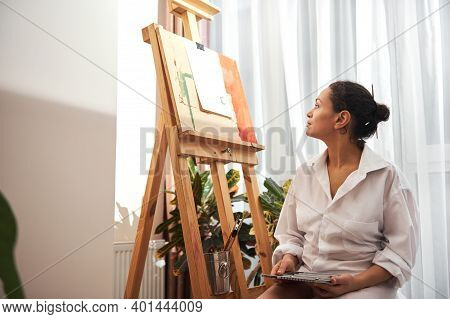 Woman Painter In White Shirt Standing In Front Of Easel Thinking And Looking At Canvas