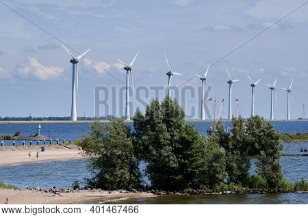 Urk, The Netherlands - June 15 2020 Windmills On The Coast With People Sunbathing And Fishing, Trees