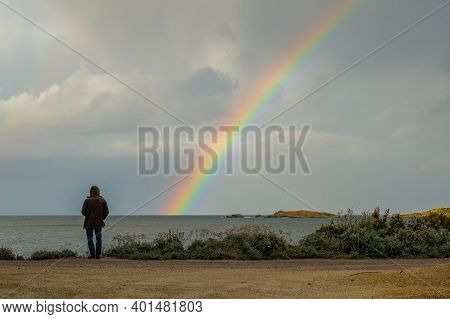 Man In Hooded Coat Looking Out Across The Mediterranean Sea Towards A Colourful Rainbow