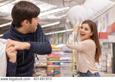 Happy Beautiful Young Woman Hitting Her Boyfriend With A Pillow, Having Fun At Furniture Store. Love