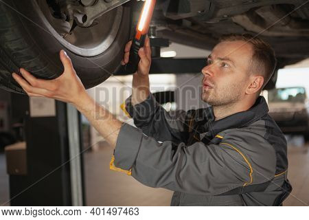 Mature Male Car Mechanic Looking Underneath A Car On The Lift, Using A Light. Experienced Auto Techn
