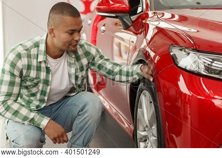 Male Customer Examining Tires Of A New Automobile On Sale. Handsome African Man Choosing New Car At 