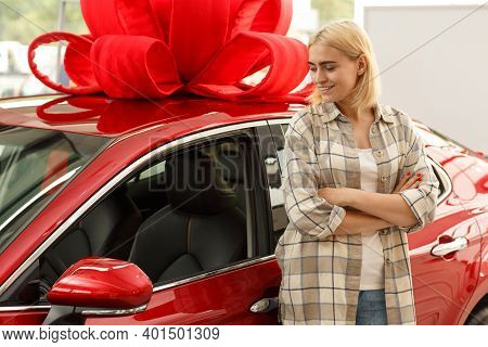 Young Attractive Woman Smiling Looking At Her New Automobile With A Big Red Bow On The Roof. Cheerfu