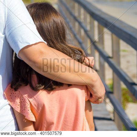 The Picture Shows A Back View Of An Unrecognizable Mother Hugging Her Daughter As They Walk