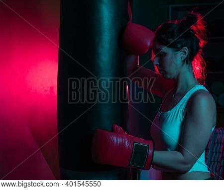 Tired Woman Boxer In Boxing Gloves Is Leaning On A Punching Bag In Red Blue Neon Light Over Dark Bac