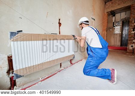 Man In Work Overalls Using Wrench While Installing Heating Radiator In Room. Young Plumber Installin