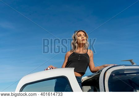 A gorgeous young blonde model poses outdoors with a single engine aircraft at a local airport