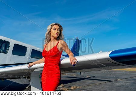 A gorgeous young blonde model poses outdoors with a single engine aircraft at a local airport
