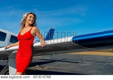 A gorgeous young blonde model poses outdoors with a single engine aircraft at a local airport