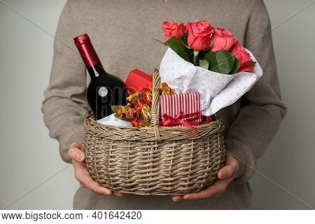 Man Holding Wicker Basket With Gifts, Bouquet And Wine On Grey Background, Closeup
