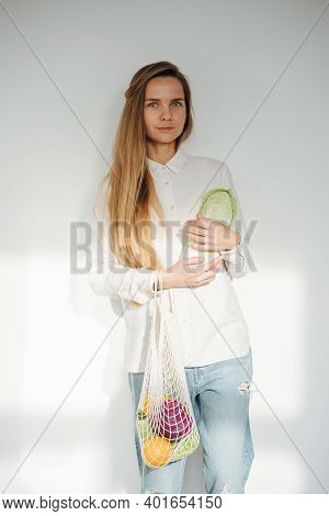 Subtely Smiling Modest Woman Posing In A Room, Holding Net Bag With Vegetables