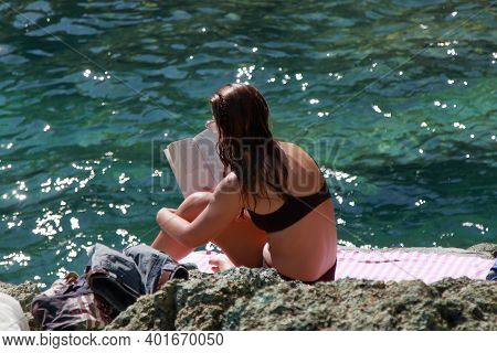 Levanto, Italy - Summer 2020: A Caucasian Woman Reads A Book Sitting On A Cliff By The Sea. High Qua