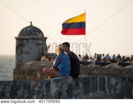 Couple Sitting On City Wall With Colombian National Flag Waving In Wind On Bastion Of Santo Domingo 