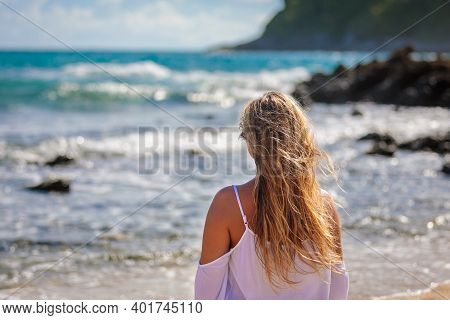 A Young Beautiful Girl Looks At The Sea, The Wind Develops Her Blond Hair. White Dress, Blue Water.,