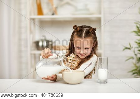 Cute Little Girl Eating Breakfast: Cereal With The Milk. Child Pours Milk Into A Bowl Of Cereal In T