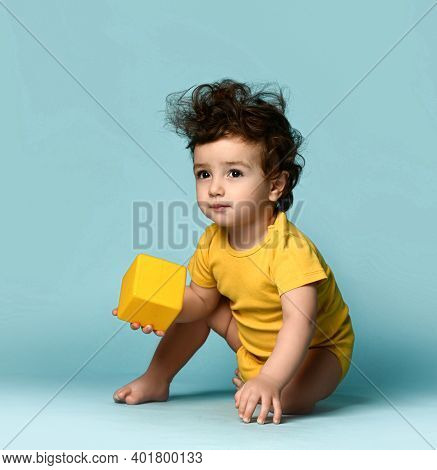 Small Cute Curly Hairy Baby Boy Toddler In Yellow Comfortable Jumpsuit Sitting On Floor And Playing 