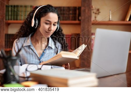 Portrait Of Calm Young Woman Wearing Wireless Headset, Reading Paper Book, Sitting At Table With Lap