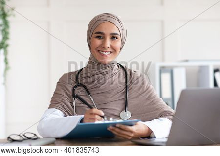 Beautiful Muslim Woman Doctor Writing In Medical Chart And Smiling At Camera, Wearing Hijab, Sitting