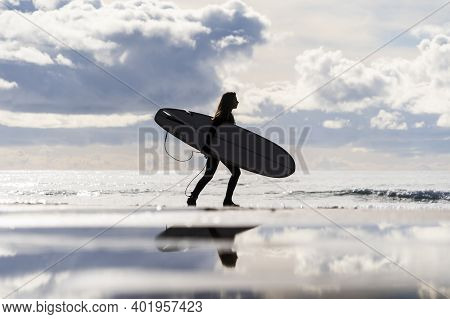 Young Surfer Girl Silhouette In Wetsuit Walking On The Beach Towards The Ocean To Catch Waves And Su