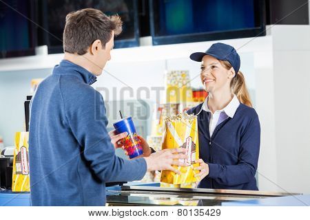 Smiling female seller giving popcorn paperbag and drink to man at concession stand in cinema