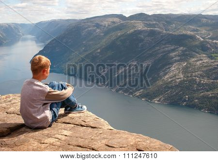 The Boy Thoughtfully Looks Afar, Sitting On The Edge Of Pulpit Rock In Norway