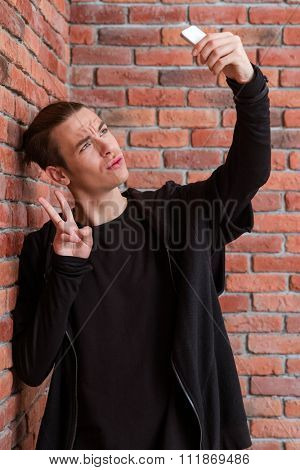 Portrait of a young man making selfie photo on smartphone oer brick wall background