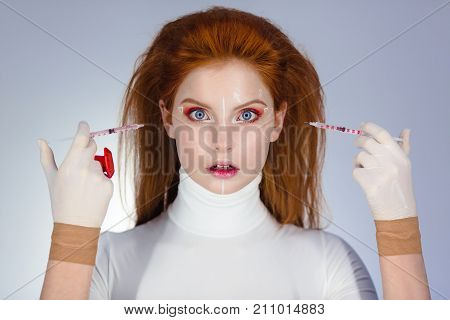Beautiful Woman before Plastic Surgery Operation Cosmetology. Beauty Portrait Fashion Concept. Treatment with botox or hyaluronic collagen injection. Young red hair girl holding syringes in hands pointing on her scared face with correction mark for plasti