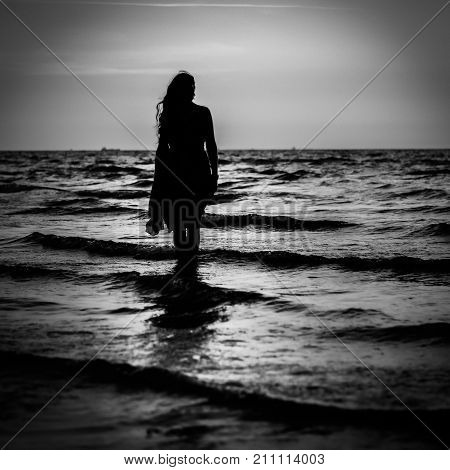 Black and white sunset portrait. Silhouette of a beautiful woman on the beach in a summer sunset. A young lady stands in the water and enjoys nature. Colorful view.