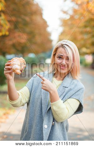 A girl in a gray jacket holds a croissant in her hand in the park shows her finger on it